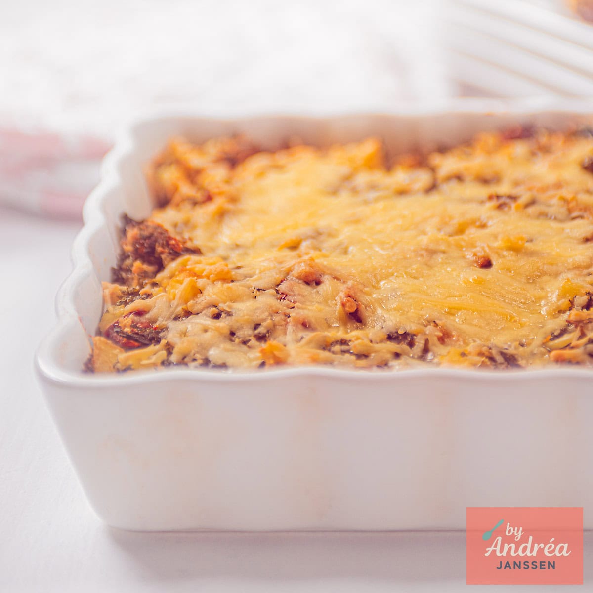 Een vierkante foto met een deel van een witte ovenschaal gevuld met een gratin van boerenkoolstamppot, kaas, en tomaat