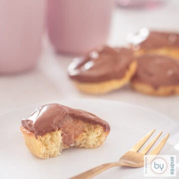 Een vierkante foto met een witte ondergrond met een wit bord met een halve cupcake a la Twix met karamel en chocola. In de achtergrond een stapeltje cupcakes