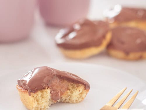 Een vierkante foto met een witte ondergrond met een wit bord met een halve cupcake a la Twix met karamel en chocola. In de achtergrond een stapeltje cupcakes