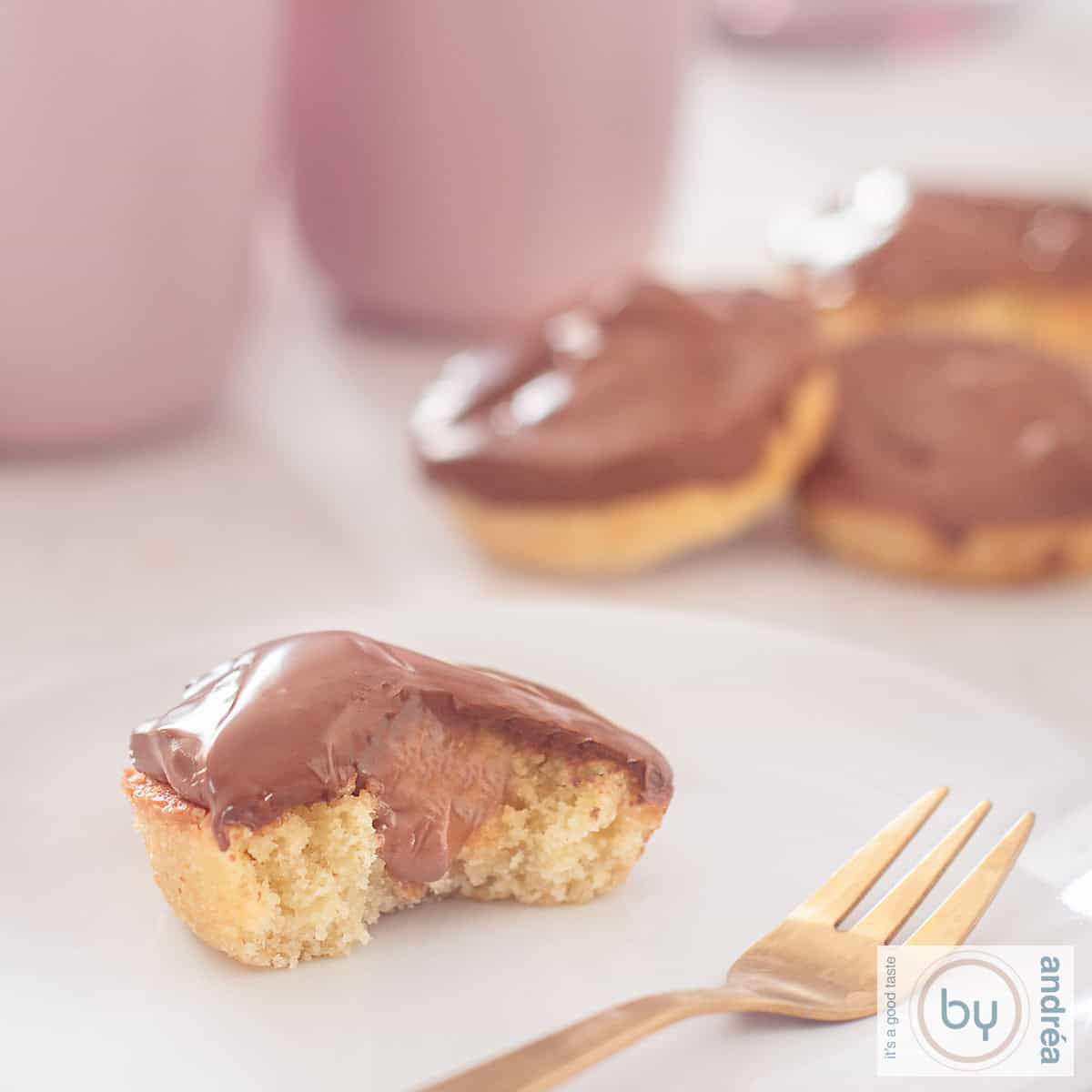 Een vierkante foto met een witte ondergrond met een wit bord met een halve cupcake a la Twix met karamel en chocola. In de achtergrond een stapeltje cupcakes