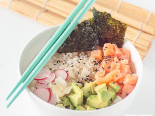 Een vierkante foto met een witte kom met sushi rijst, radijs, avocado, gerookte zalm en zeewier. Twee blauwe eetstokjes en een rieten mat kleden het aand