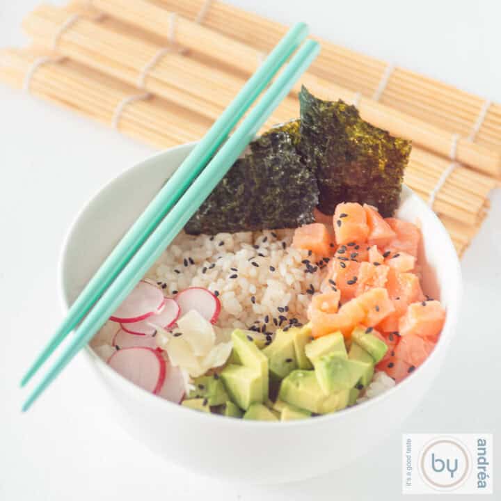 Een vierkante foto met een witte kom met sushi rijst, radijs, avocado, gerookte zalm en zeewier. Twee blauwe eetstokjes en een rieten mat kleden het aand
