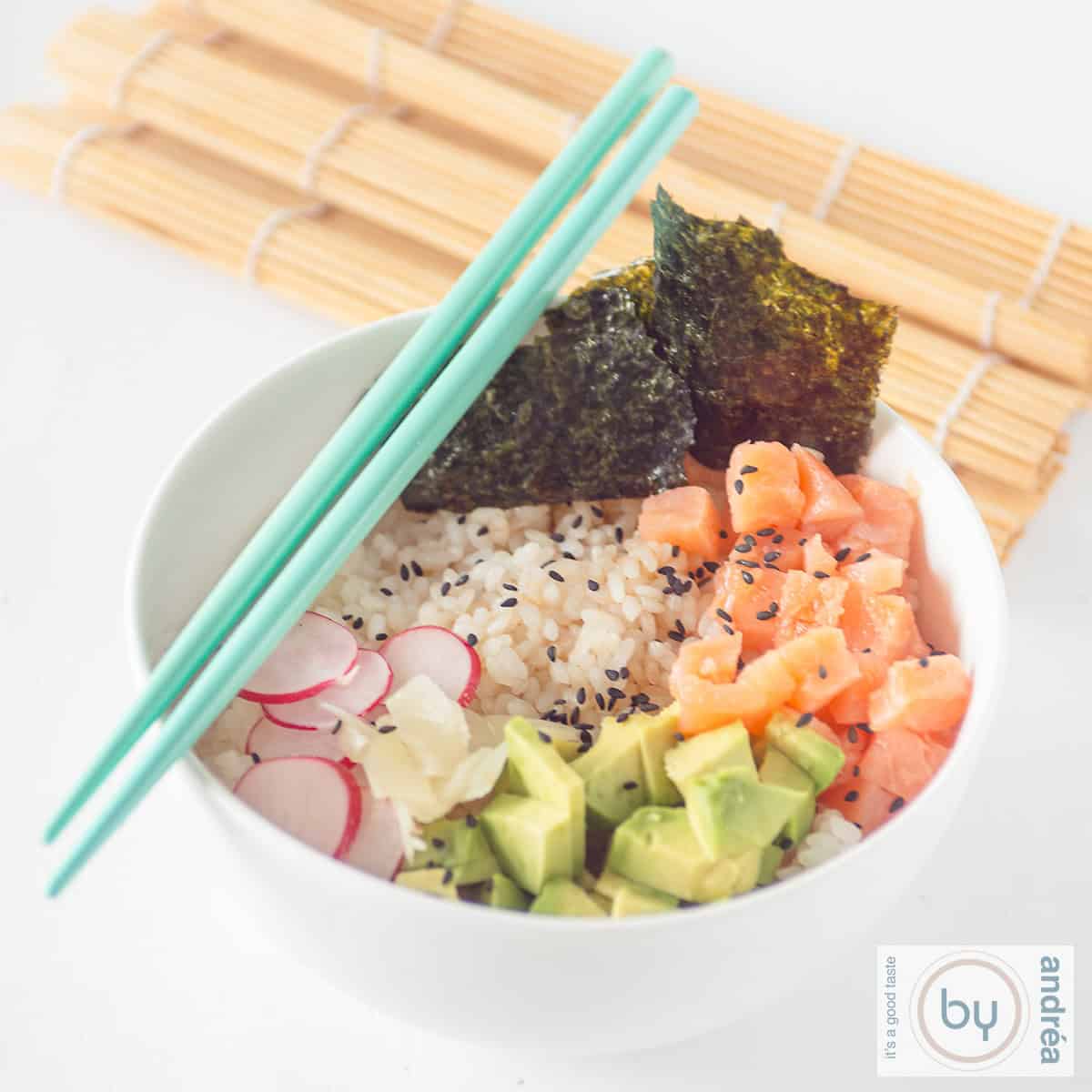 Een vierkante foto met een witte kom met sushi rijst, radijs, avocado, gerookte zalm en zeewier. Twee blauwe eetstokjes en een rieten mat kleden het aand