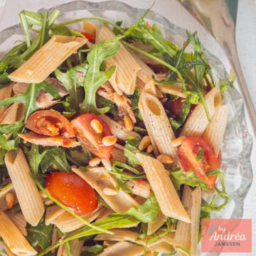 Een vierkante foto van een glazen schaal met penne, tomaatjes, rucola, makreel en tapenade