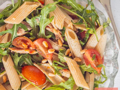 Een vierkante foto van een glazen schaal met penne, tomaatjes, rucola, makreel en tapenade