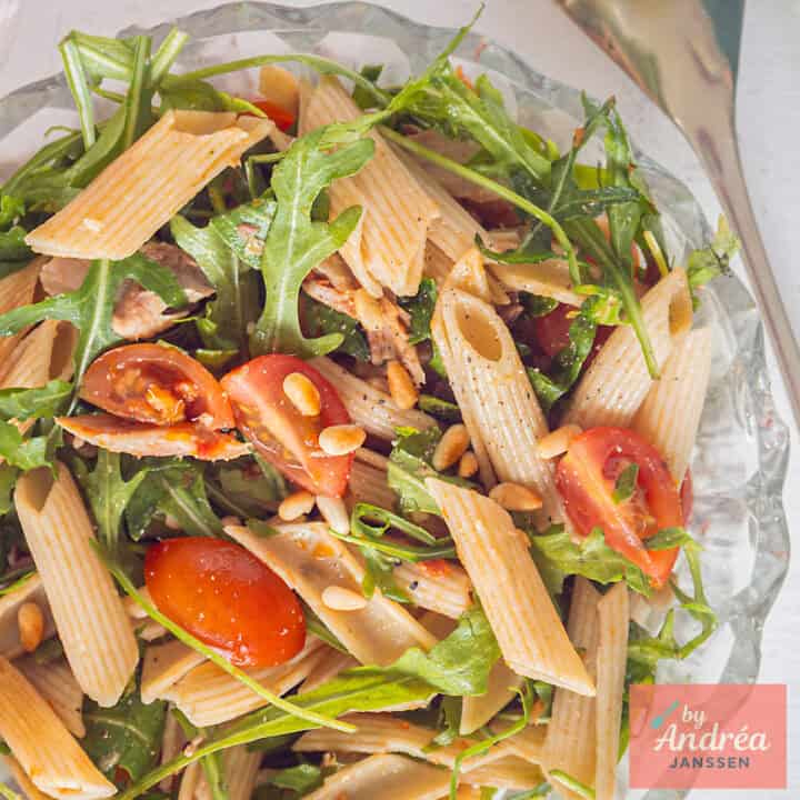Een vierkante foto van een glazen schaal met penne, tomaatjes, rucola, makreel en tapenade