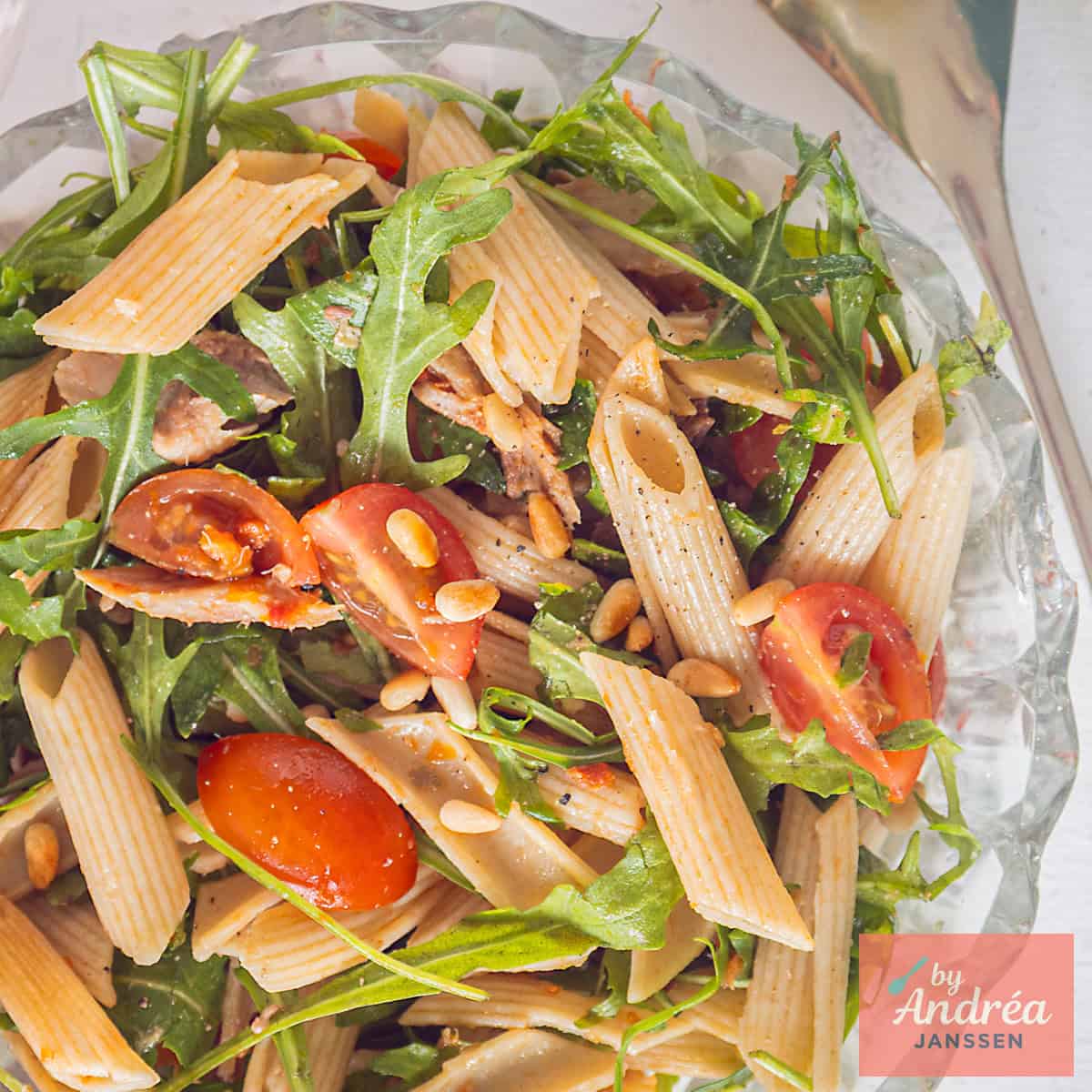Een vierkante foto van een glazen schaal met penne, tomaatjes, rucola, makreel en tapenade