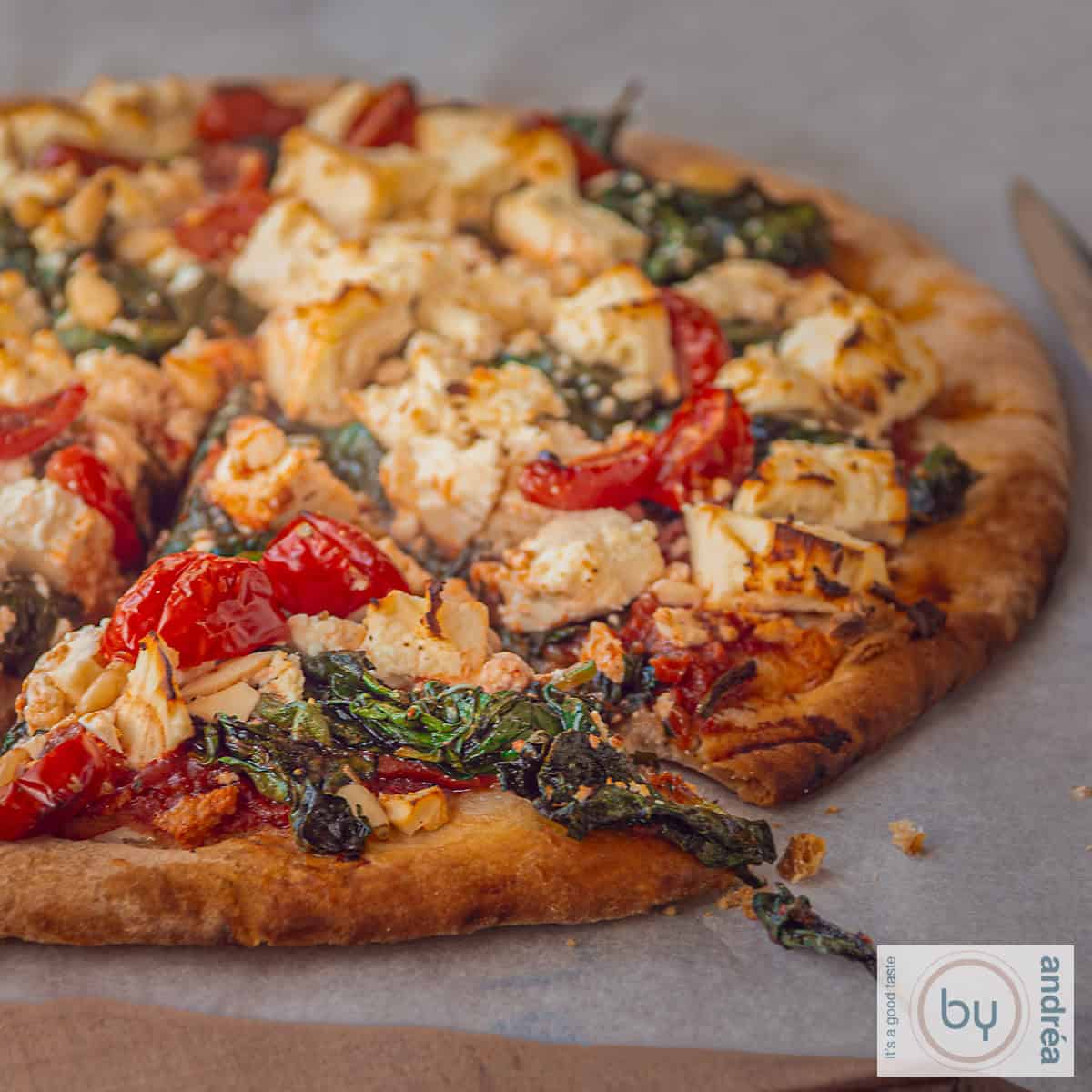 Een vierkante foto met een pizza, waaruit een punt is gesneden op een wit bakpapier. De pizza is belegt met feta, tomaat, spinazie en pijnboompitjes