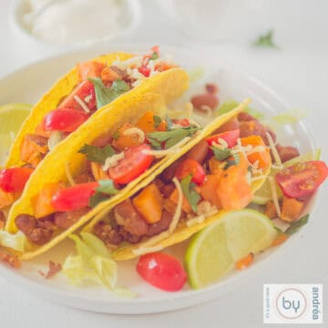 Een vierkante foto van een wit bord met drie gevulde taco's met zoete aardappel, kaas en bonen. Een heerlijk vegetarisch hoofdgerecht