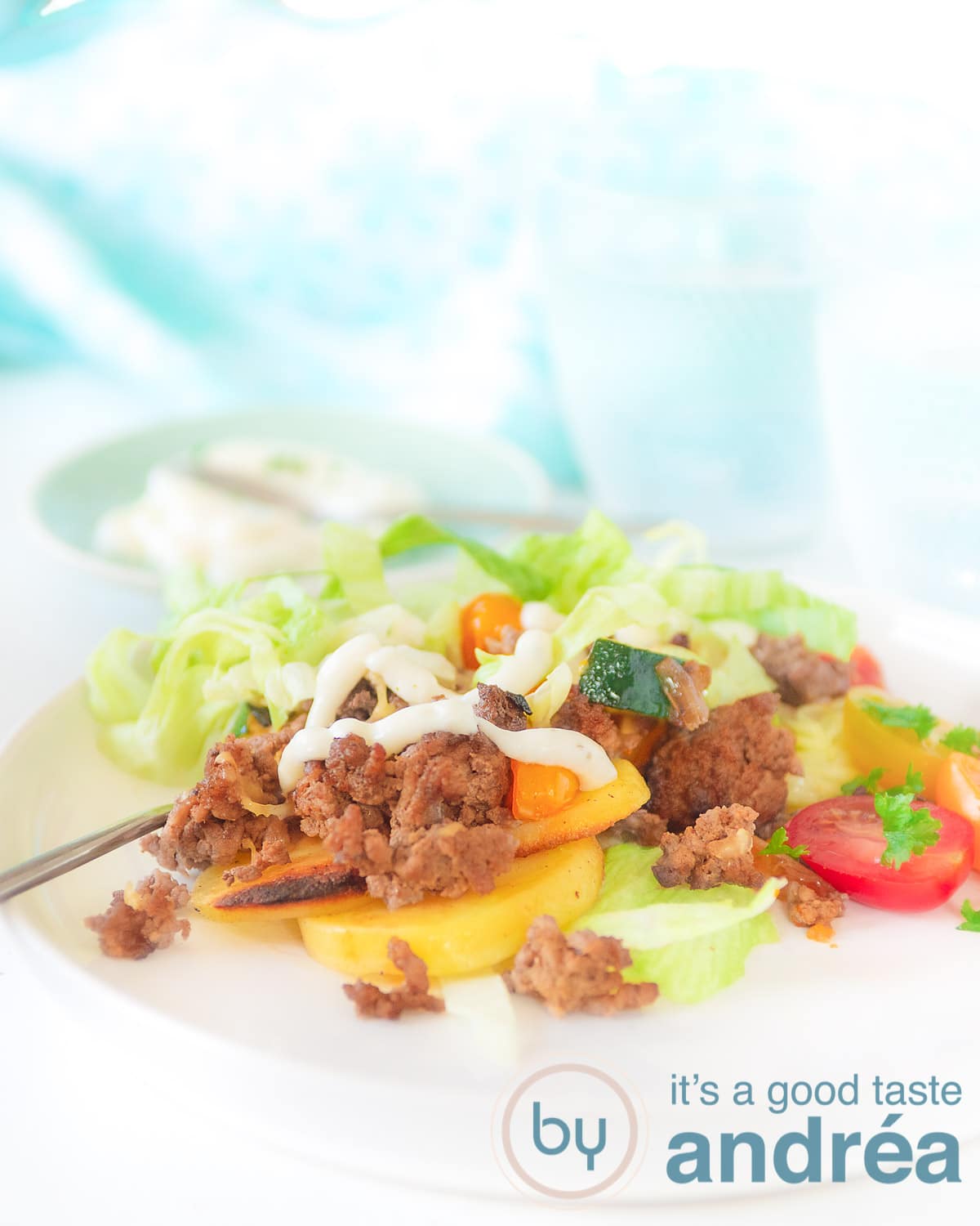 Een wit bord met aardappeltjes, gehakt, ijsbergsla gegarneerd met tomaat en knoflooksaus