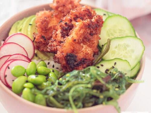 Een vierkante foto met een roze kom: een sushibowl gevuld met sriracha kip, zeewiersalade, edamame bonen, radijs en komkommer