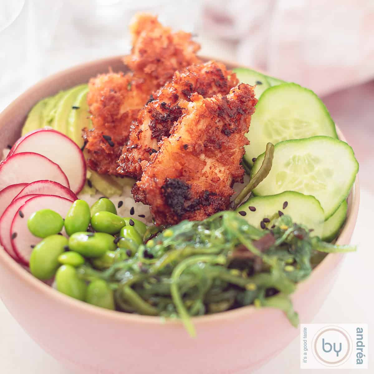 Een vierkante foto met een roze kom: een sushibowl gevuld met sriracha kip, zeewiersalade, edamame bonen, radijs en komkommer