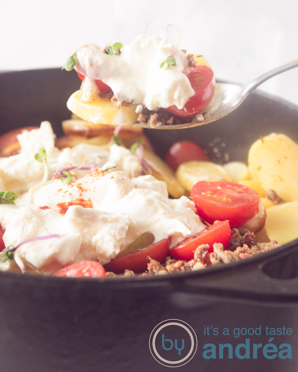 Greek potato dish with minced meat in a black pan. A spoon takes out a scoop.