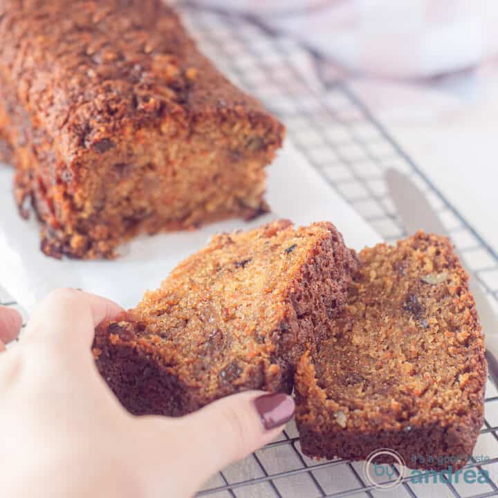 Een vierkante foto met een cake met wortels, walnoten en rozijnen op een bakpapier op een rek. Twee plakjes erafgesneden. Een hand pakt het bovenste plakje.