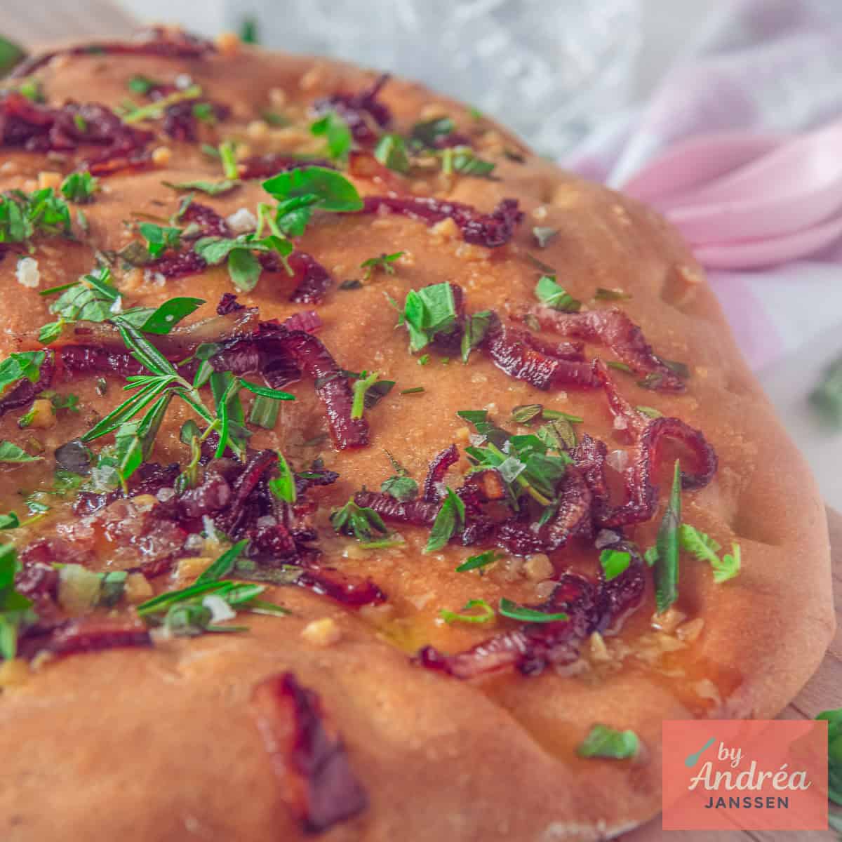 Een vierkante foto met een deel van een focaccia met een topping van verse kruiden, boter, knoflook en verse kruiden.