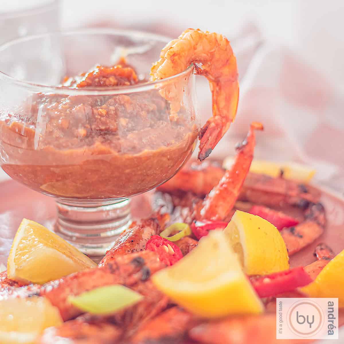 Een vierkante foto met een glas gevuld met cashew satésaus en gegrilde garnalen, bosui, citroen en pepers eromheen.