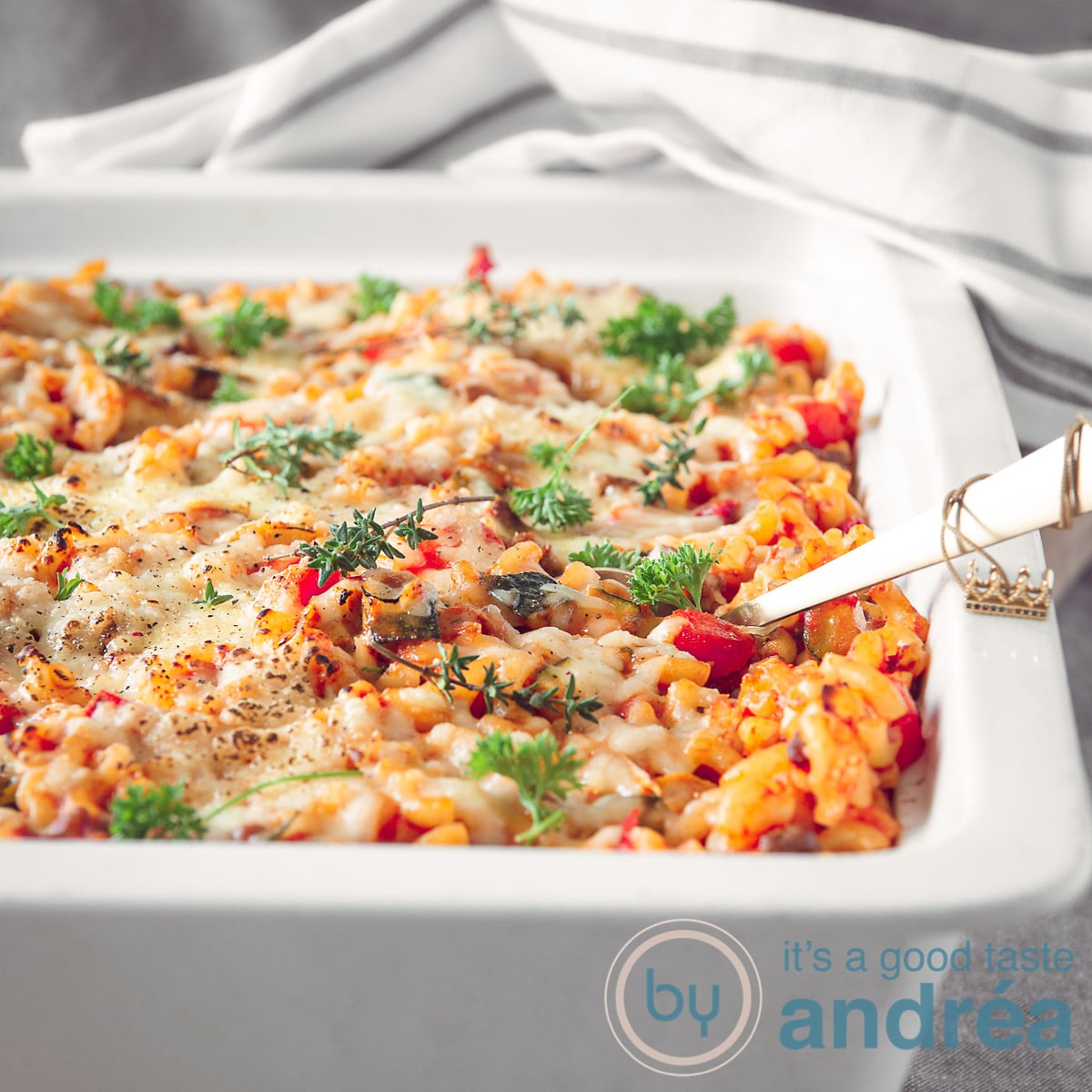 Een macaroni ovenschotel in een witte schaal. Op de achtergrond een grijs wit gestreepte handdoek