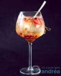 A black background with a glass filled with sangria with fresh summer fruit and licor 43. A golden spoon stand inside the glass