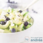 Een vierkante foto met een deel van een witte schaal gevuld met meloensalade met blauwe bessen en feta en komkommer op een witte achtergrond.
