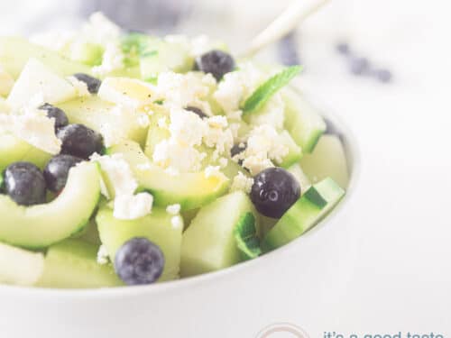 Een vierkante foto met een deel van een witte schaal gevuld met meloensalade met blauwe bessen en feta en komkommer op een witte achtergrond.