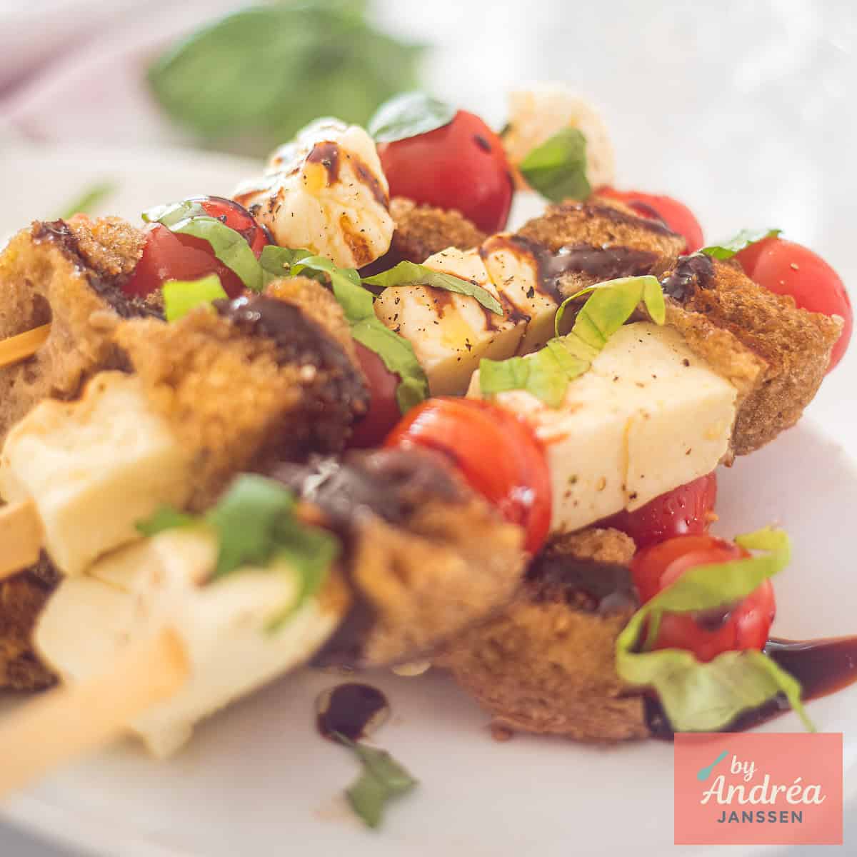 Een vierkante foto van een bord met spiesjes met halloumi, cherrytomaten, knoflookbrood, basilicum en balsamicocreme