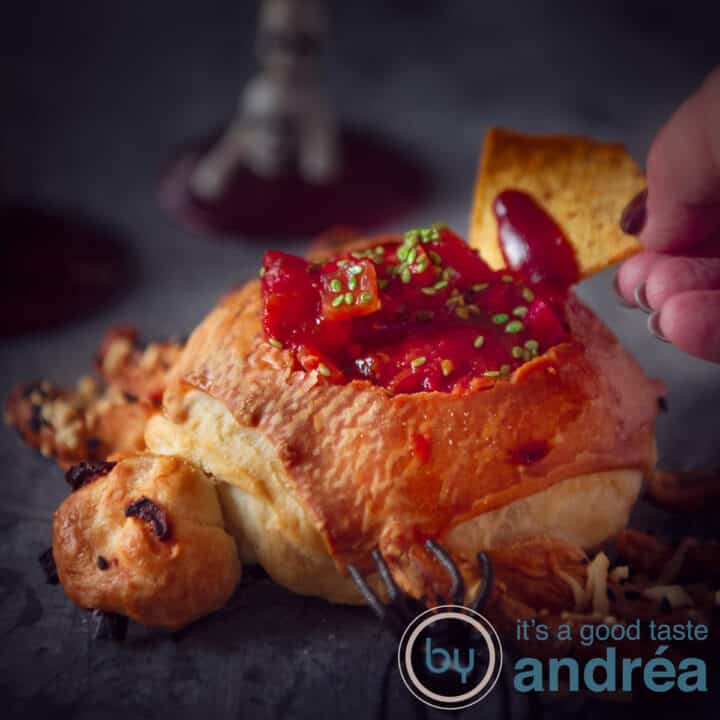 A square picture of a bread bowl in the form of a spider, containing taco sauce. A nacho dips in the sauce. A dark background