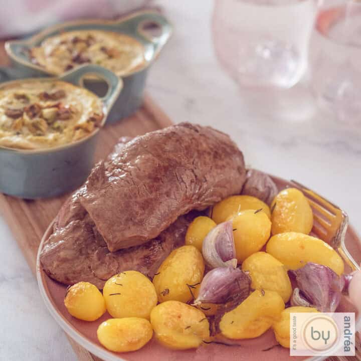 Een vierkante foto met een roze bord met krieltjes, gepofte knoflook en lendebiefstuk. Er echter twee schaaltjes met broccoli geitenkaas quiche