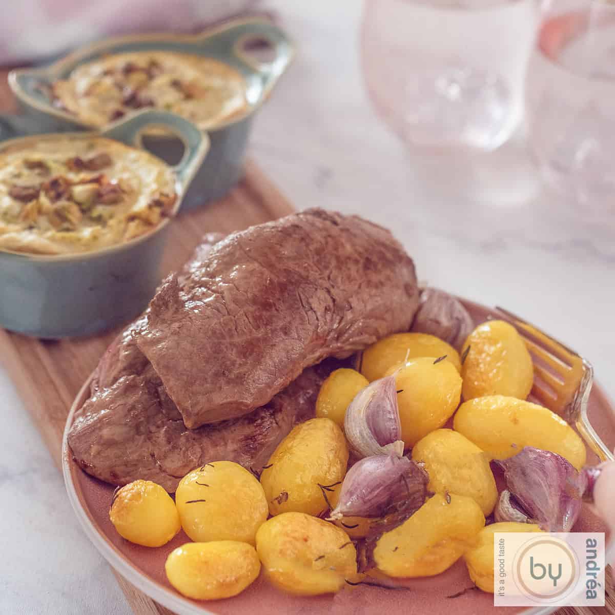 Een vierkante foto met een roze bord met krieltjes, gepofte knoflook en lendebiefstuk. Er echter twee schaaltjes met broccoli geitenkaas quiche