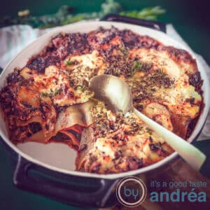 Een vierkante foto met een zwarte ovenschaal gevuld met lasagne van boerenkool en worst. Een klein deel is eruit. Een gouden lepel steekt in de lasagne.