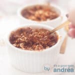 Een vierkante foto van twee ramekins gevuld met appeltaart granola. Een hand pakt een lepel eruit.