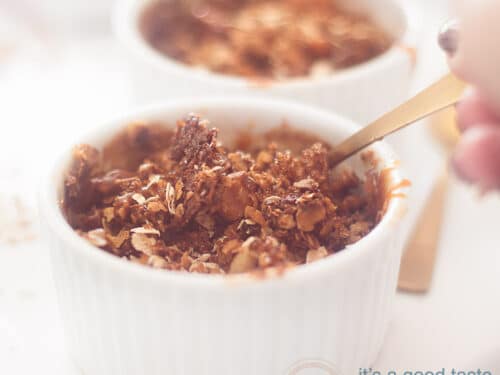 Een vierkante foto van twee ramekins gevuld met appeltaart granola. Een hand pakt een lepel eruit.