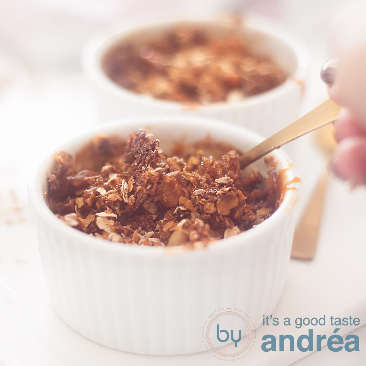 Een vierkante foto van twee ramekins gevuld met appeltaart granola. Een hand pakt een lepel eruit.