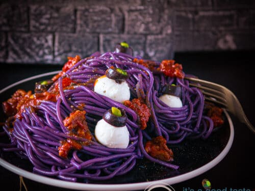 Een vierkante foto met een zwart bord op een donkere ondergrond, paarse spaghetti, oogballen van mozzarella en tomatensaus