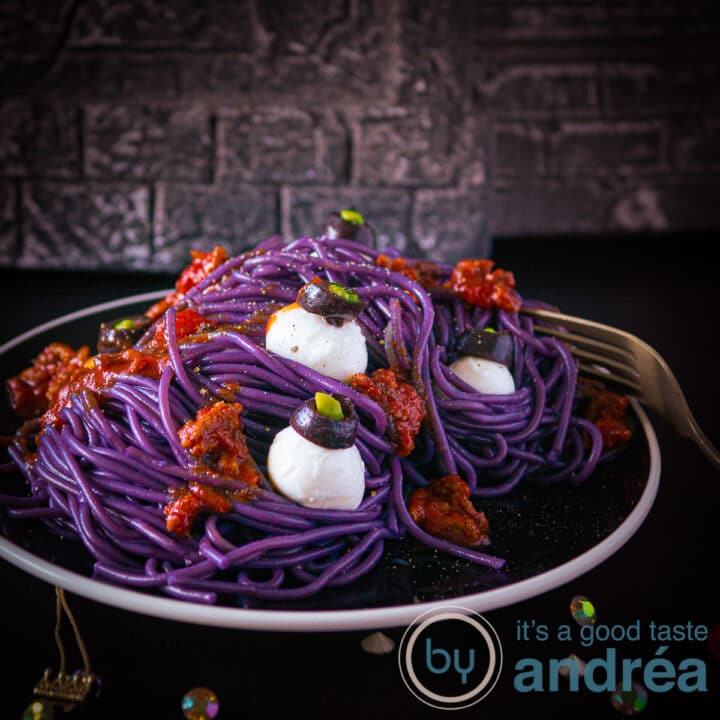 Een vierkante foto met een zwart bord op een donkere ondergrond, paarse spaghetti, oogballen van mozzarella en tomatensaus