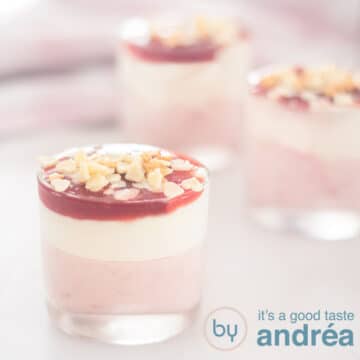 Een vierkante foto met drie dessert glaasjes gevuld met aardbeien met witte chocolade mousse en een glazuur. Een roze witte handdoek in de achtergrond