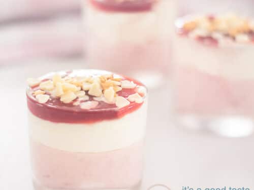 Een vierkante foto met drie dessert glaasjes gevuld met aardbeien met witte chocolade mousse en een glazuur. Een roze witte handdoek in de achtergrond