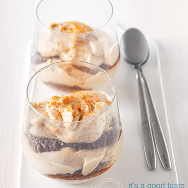 Twee glazen gevuld met Speculoos mascarpone room afgewisseld met koek op een wit plaatje achter elkaar. Twee lepels aan de zijkant. Een witte ondergrond