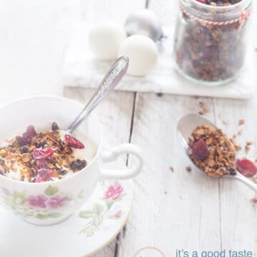 Een kopje gevuld met muesli, cranberries. Kerstballen en een potje met granola in de achtergrond. Een lepel met granola in de rechterkant.