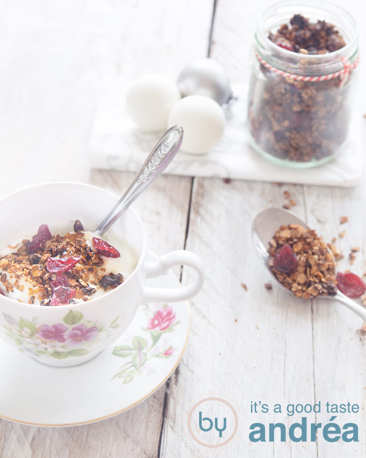 Een kopje gevuld met muesli, cranberries. Kerstballen en een potje met granola in de achtergrond. Een lepel met granola in de rechterkant.