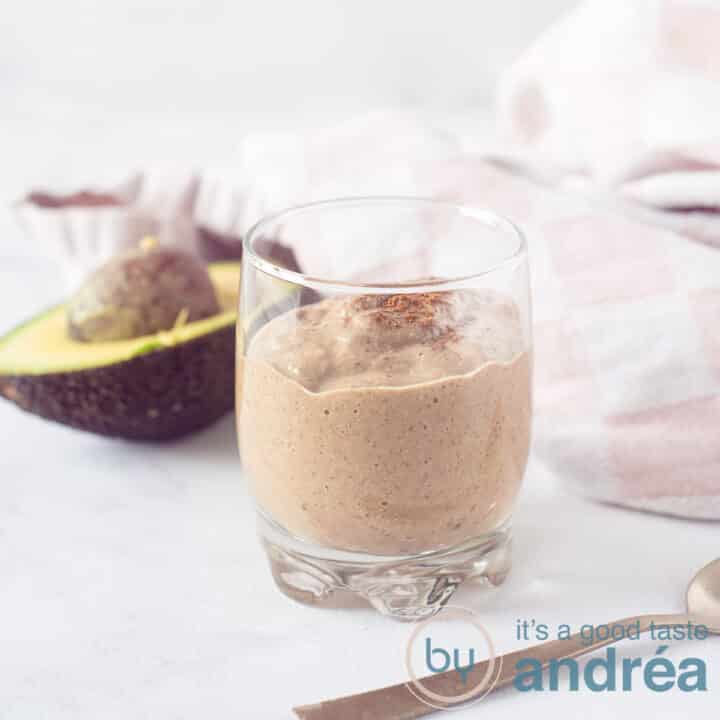 Een vierkante foto met een glas met chocolade kokos smoothie. Een avocado en een roze witte theedoek in de achtergrond.