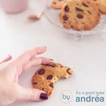 Een vierkante foto met een half koekje dat een hand vastpakt. Een witte ondergrond. Rechtsbovenin een schaal met chocolate chip koekjes.