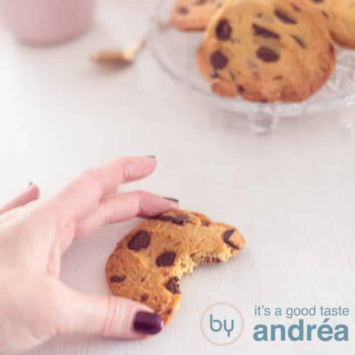 Een vierkante foto met een half koekje dat een hand vastpakt. Een witte ondergrond. Rechtsbovenin een schaal met chocolate chip koekjes.