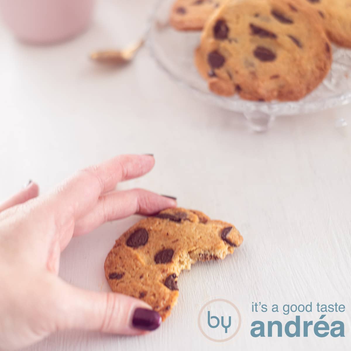 Een vierkante foto met een half koekje dat een hand vastpakt. Een witte ondergrond. Rechtsbovenin een schaal met chocolate chip koekjes.