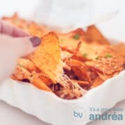 Een vierkante foto met een witte ovenschotel gevuld met nacho ovenschotel met gehakt en kaas. Een hand pakt een nacho chip en trekt hem los. Een witte achtergrond.