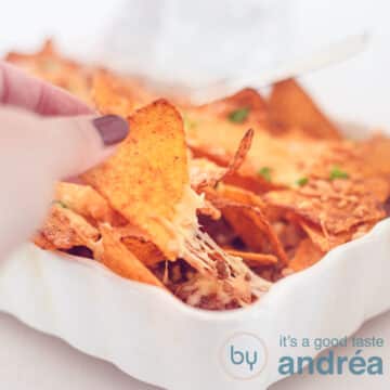 Een vierkante foto met een witte ovenschotel gevuld met nacho ovenschotel met gehakt en kaas. Een hand pakt een nacho chip en trekt hem los. Een witte achtergrond.