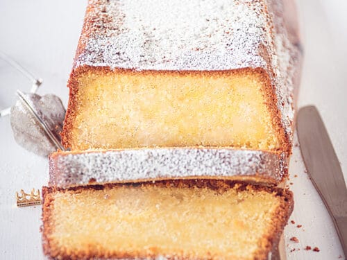 Een vierkante foto met een houten plank met daarop een cake gemaakt met roomkaas en zure room. Twee plakjes erafgesneden. Een witte ondergrond.