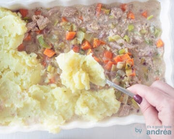 Aardappelpuree wordt over de groentevulling met tonijn geschept in een witte ovenschaal.