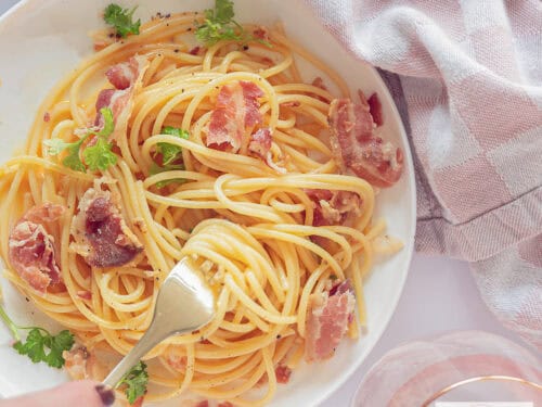 Een vierkante foto van bovenaf genomen vanaf een wit bord zonder pasta carbonara. Een vork prikt in de spaghetti
