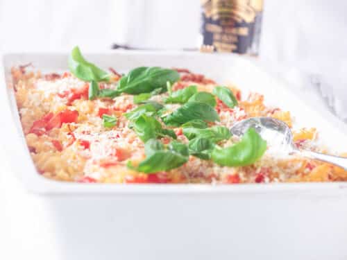 Een vierkante foto met een witte ovenschaal gevuld met een pasta ovenschotel met tomaten, mascarpone en basilicum. Een witte ondergrond.