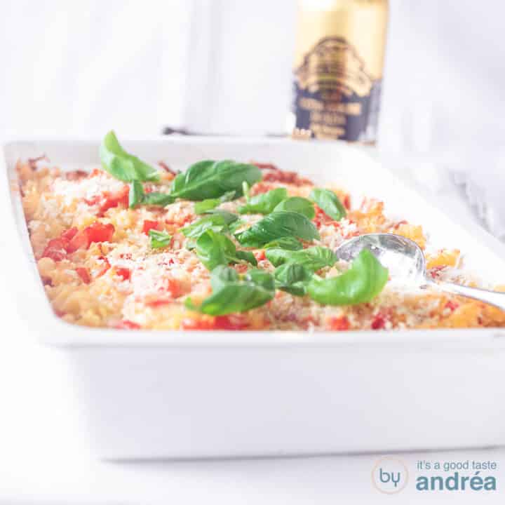 Een vierkante foto met een witte ovenschaal gevuld met een pasta ovenschotel met tomaten, mascarpone en basilicum. Een witte ondergrond.